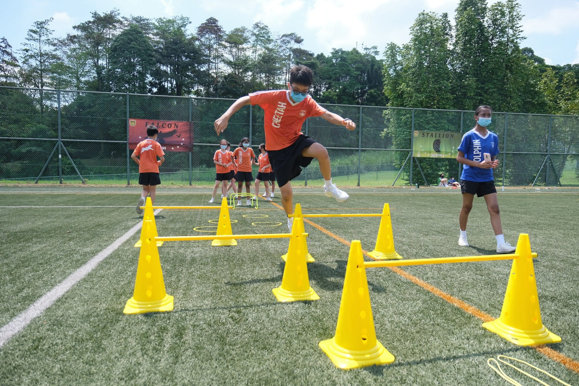 A masked boy in an orange shirt leaps over yellow cone hurdles on a green turf field with other masked students watching.